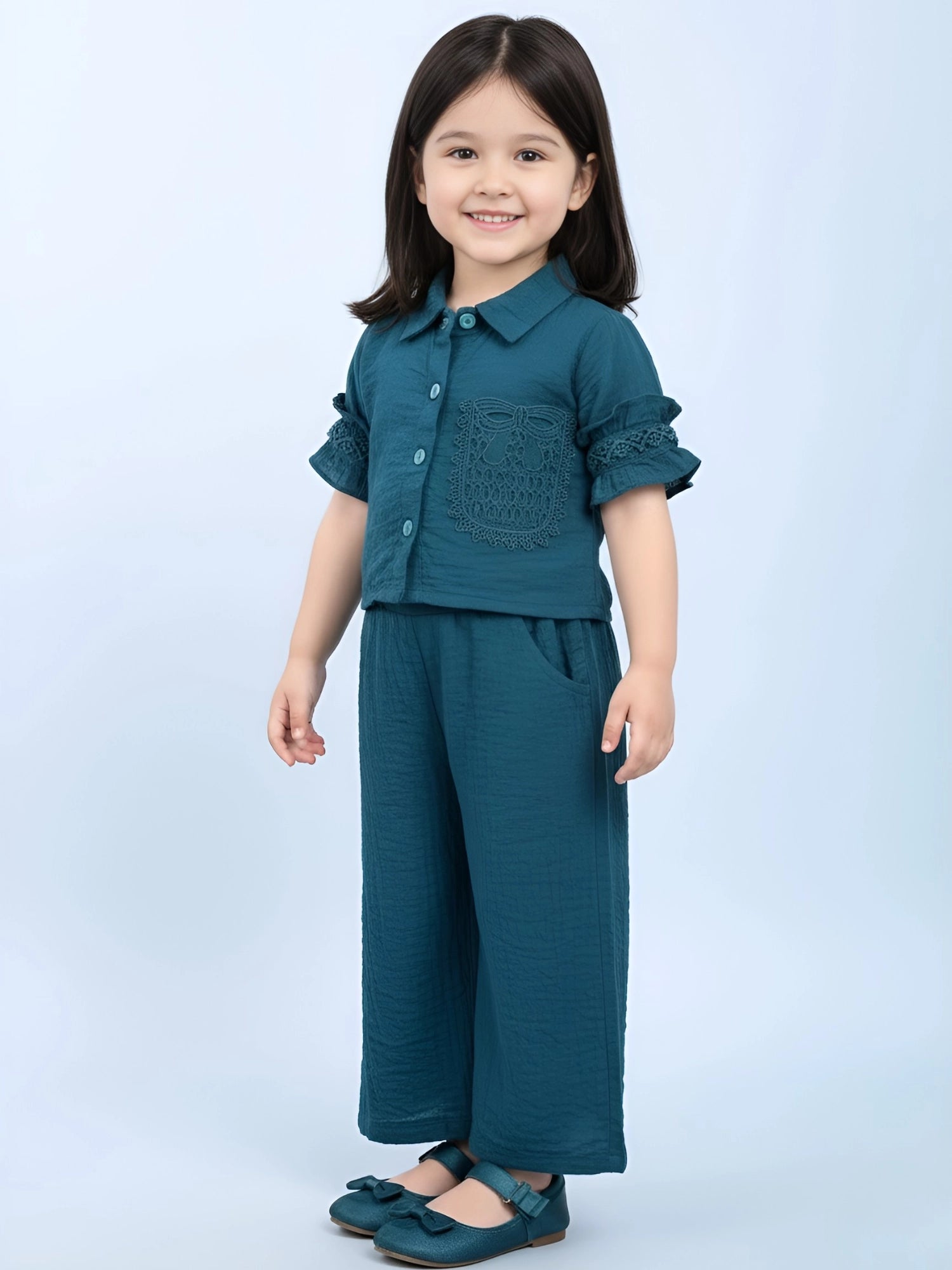 Little girl wearing a two-piece teal textured co-ord set with ruffle sleeves, button-down shirt, and wide-leg trousers against a light blue background