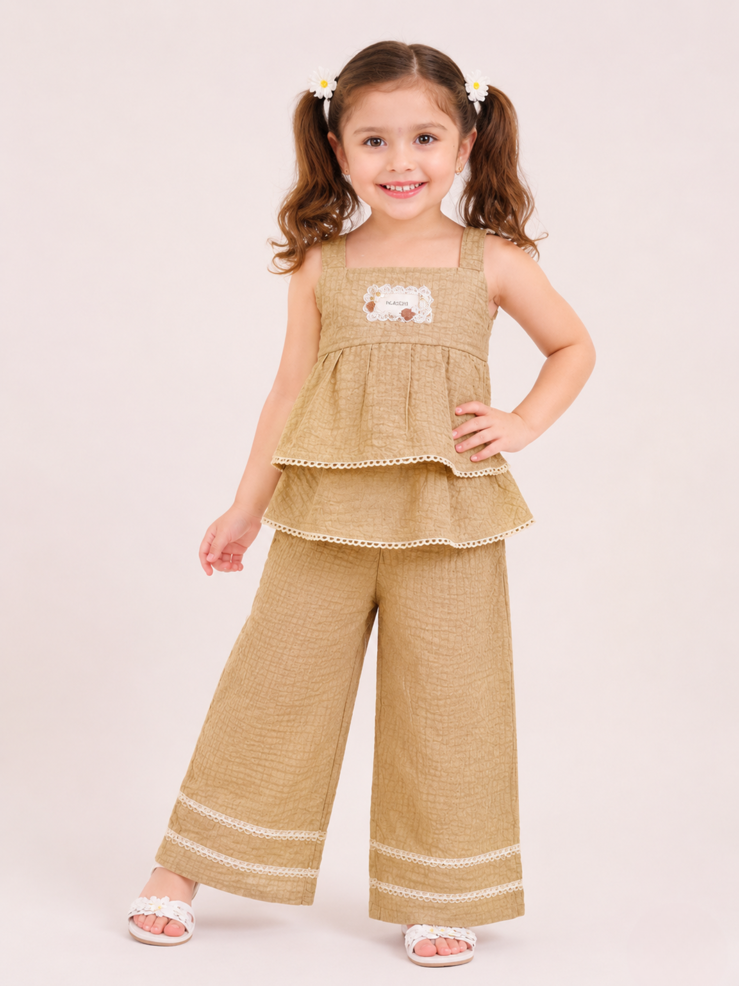 A young girl posing with one hand on her hip in a studio setting wearing a two-piece co-ord set from Kiya Store, featuring a beige textured sleeveless layered peplum top with white lace edges, paired with matching wide-leg pants decorated with dual lace borders near the hem.