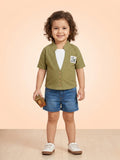 A toddler girl with curly hair wearing an olive green ribbed short-sleeve waistcoat over a white tee, paired with denim shorts and white sneakers.