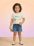 A smiling toddler girl with curly hair wearing a cream-colored short-sleeve tee with "SHINE FREEDOM" and floral illustrations, paired with denim shorts and a small bunny toy.