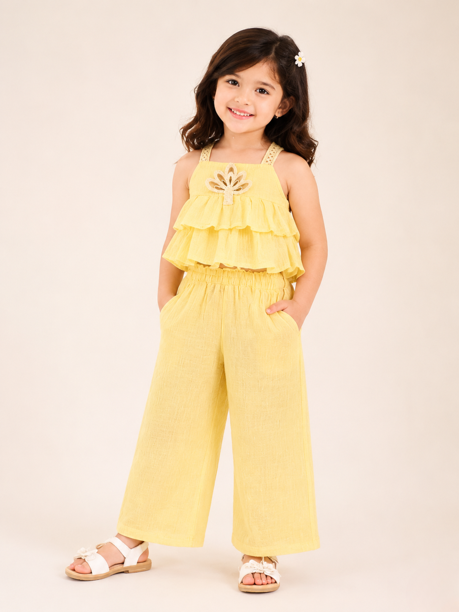 A young girl posing with her hands in her pockets in a studio setting wearing a two-piece co-ord set from Kiya Store, featuring a pastel yellow sleeveless layered crop top with a crown-like geometric cutout motif on the chest, paired with matching loose wide-leg trousers.