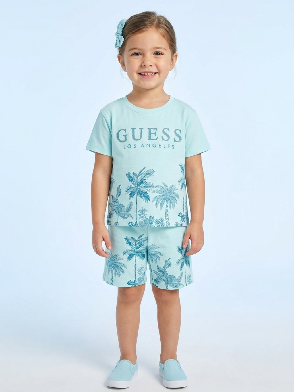 a young girl wears a mint green short-sleeve t-shirt with "GUESS Los Angeles" branding and a blue-toned palm tree graphic, paired with matching printed shorts.