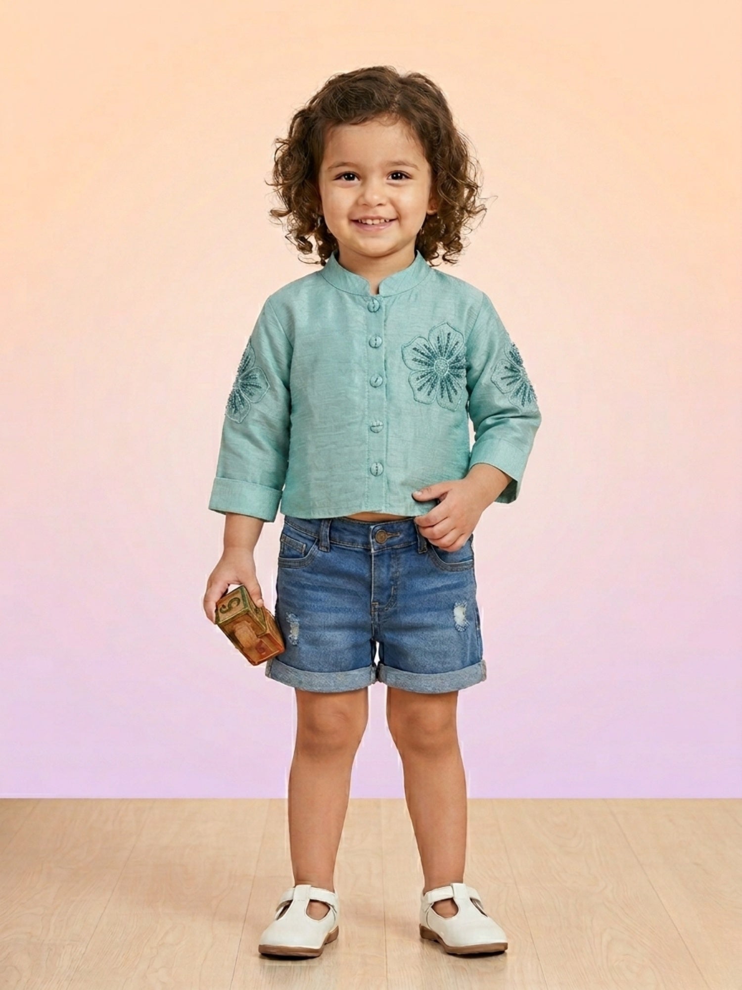 A toddler girl wearing a seafoam teal silk-textured button-up top with large tonal floral embroidery on the chest and sleeves, paired with denim shorts