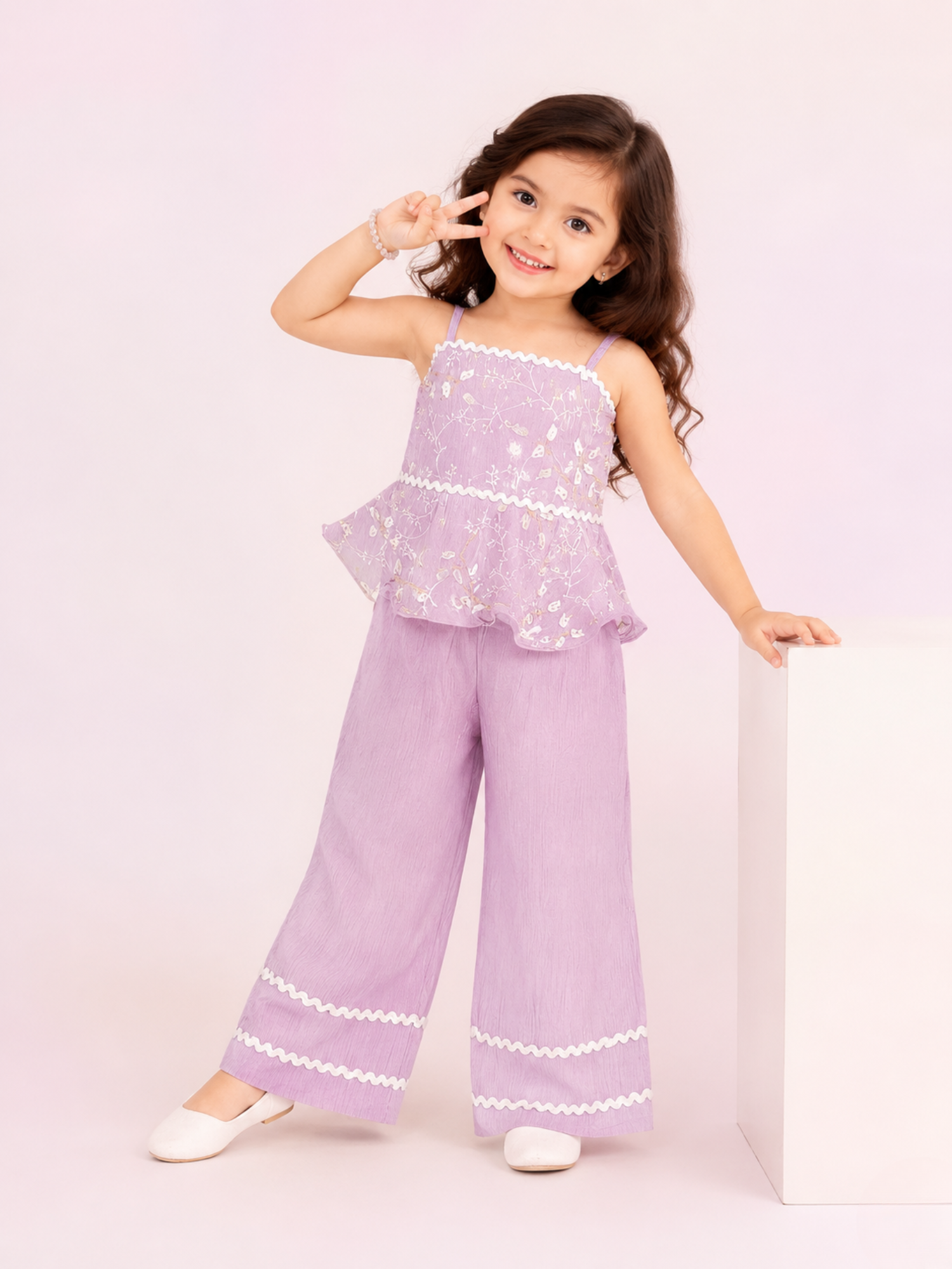 A young girl giving a peace sign while leaning on a white block in a studio setting, wearing a two-piece lavender co-ord set from Kiya Store featuring a sleeveless floral embroidered peplum top and matching wide-leg trousers with white wavy lace trim.