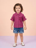 A toddler girl with curly hair wearing a magenta pink button-up shirt with ruffled sleeves and a matching floral lace appliqué on the chest, paired with denim shorts.