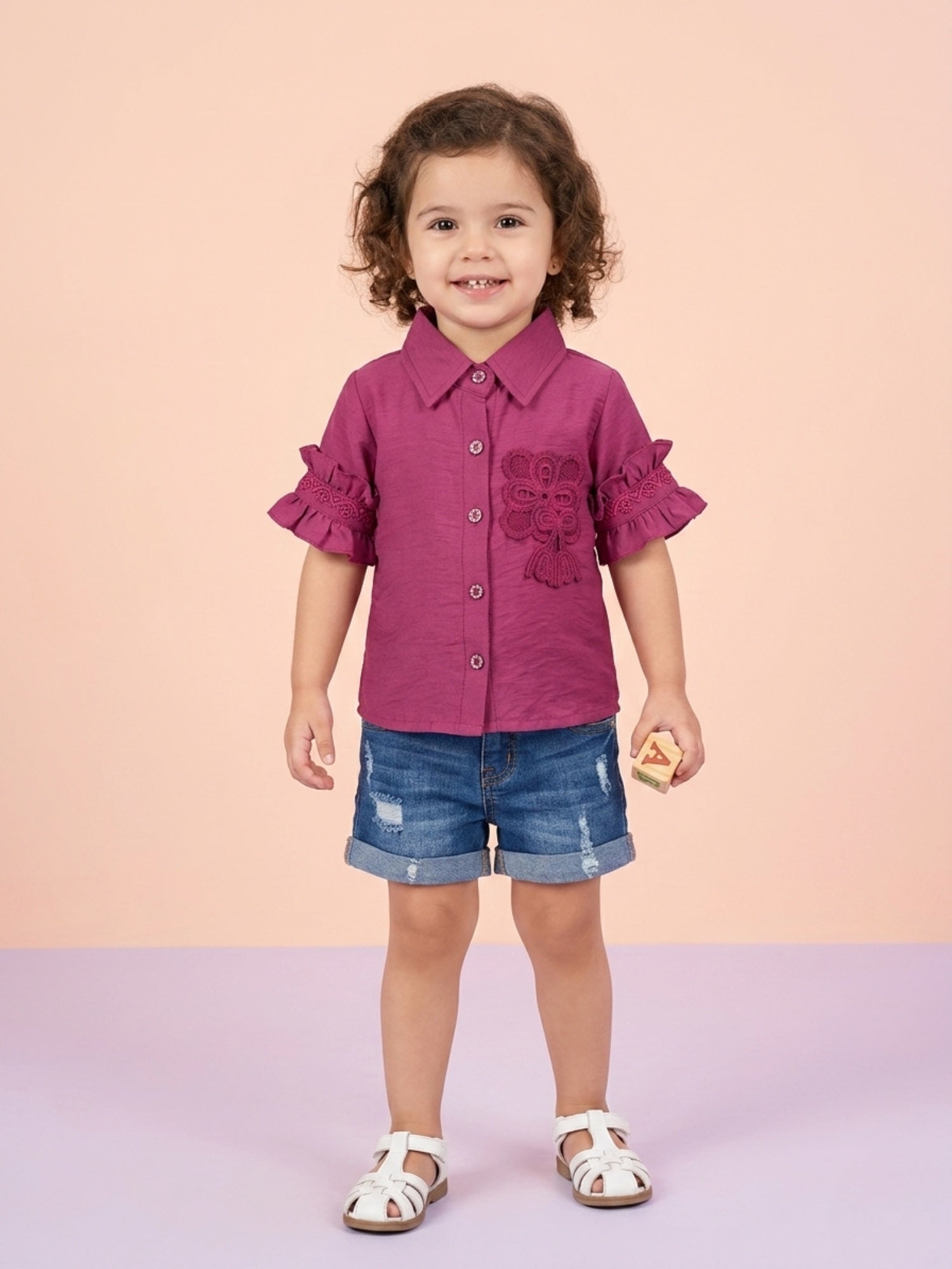 A toddler girl with curly hair wearing a magenta pink button-up shirt with ruffled sleeves and a matching floral lace appliqué on the chest, paired with denim shorts.