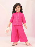 In the image titled "Untitled design(17).png", a young girl stands smiling while wearing a vibrant rani pink ethnic co-ord set, consisting of a short-sleeved top with a mandarin collar and central floral embroidery placket paired with matching wide-leg trousers. She is holding a small plush bunny toy against a soft, pastel pink background.