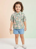 A toddler girl with curly hair wearing a sage green short-sleeve shirt with a peach floral print, utility pockets, and D-ring details, paired with light blue denim shorts.
