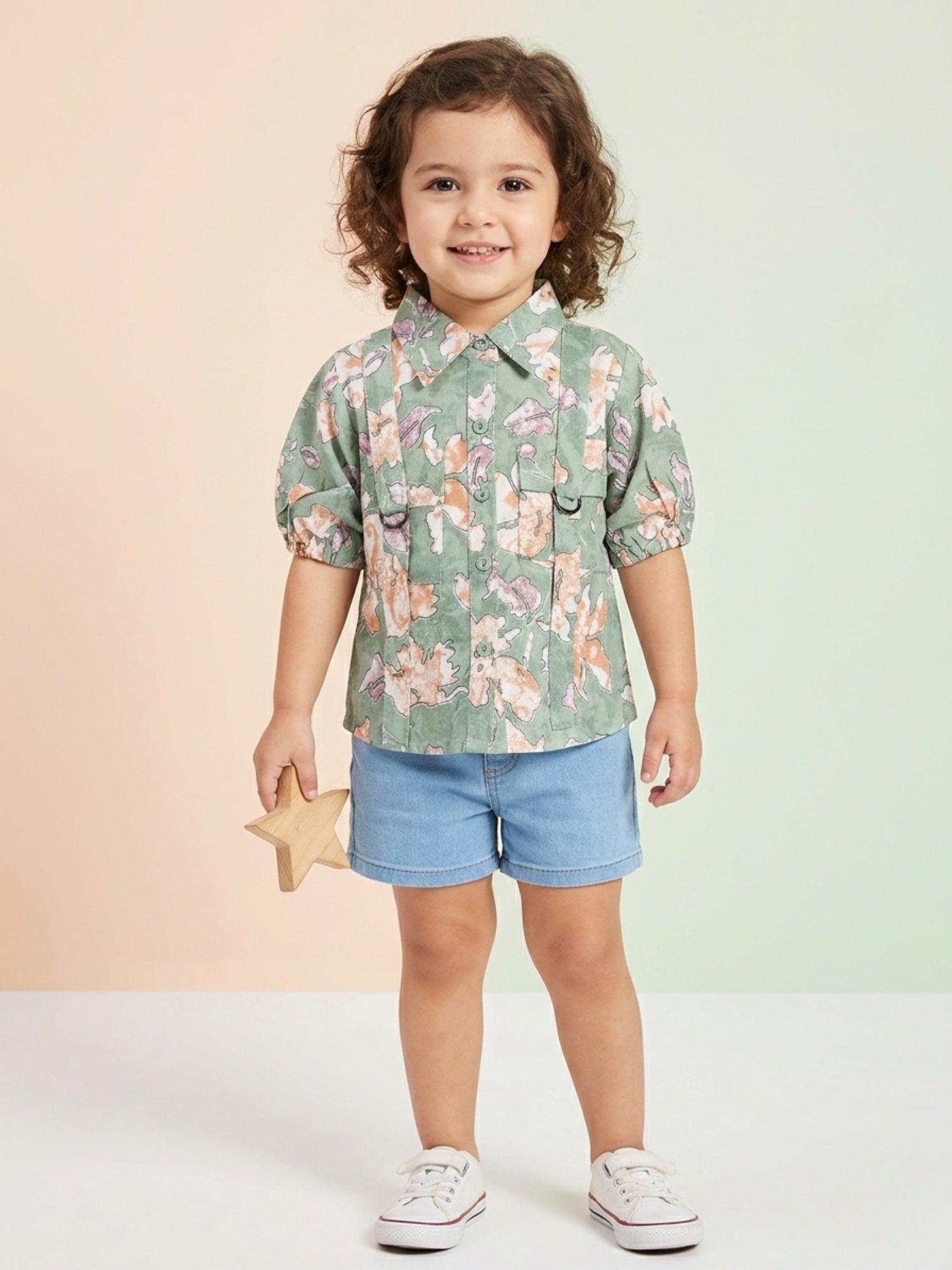 A toddler girl with curly hair wearing a sage green short-sleeve shirt with a peach floral print, utility pockets, and D-ring details, paired with light blue denim shorts.