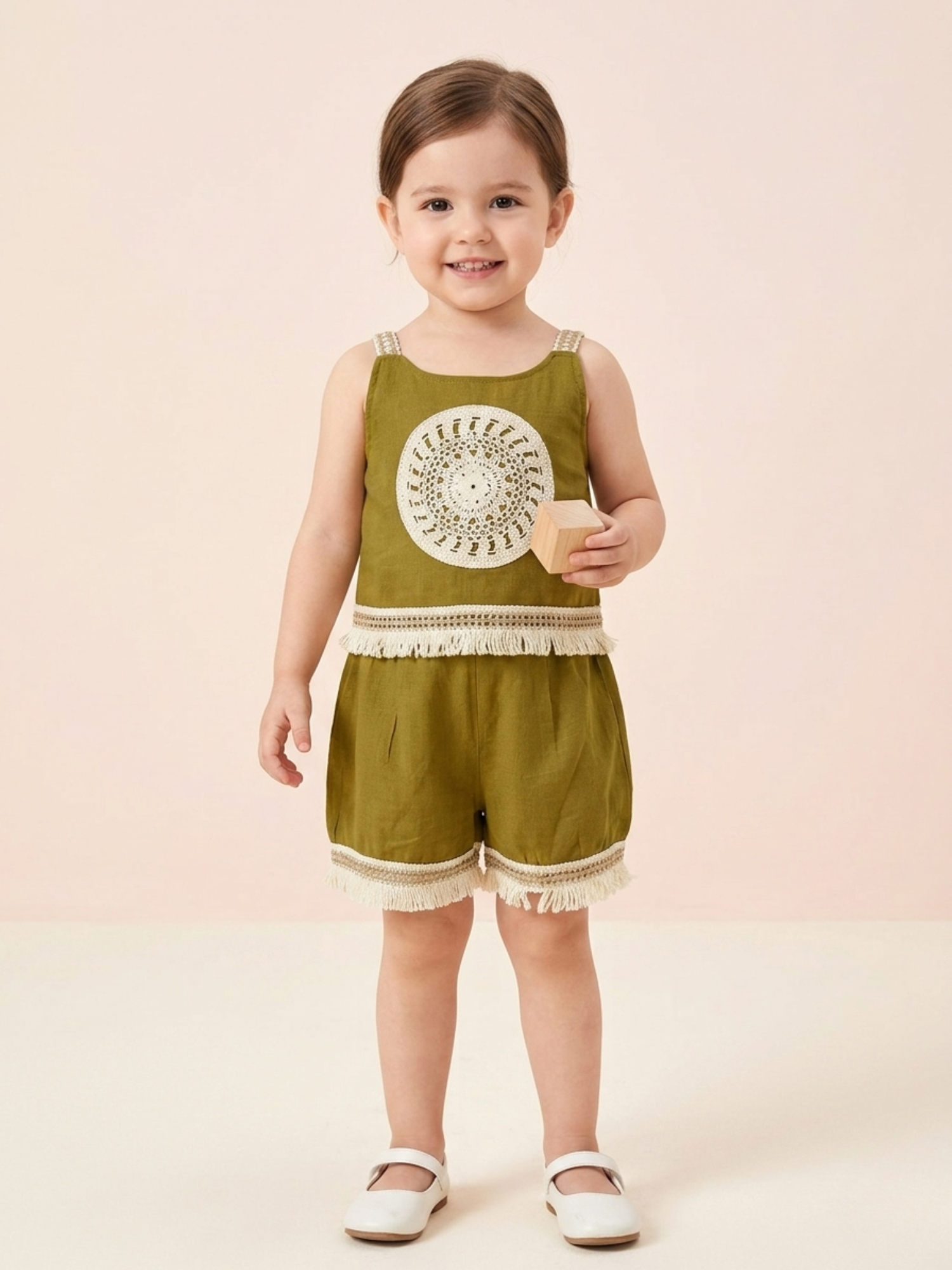 In the image titled "Untitled design(18).png", a toddler girl smiles while wearing a boho-style olive green sleeveless top and shorts co-ord set. The cami top features a cream crochet circular mandala applique on the chest with fringe lace trim at the hem, matched with coordinating fringed shorts. She holds a small wooden block against a neutral beige background.