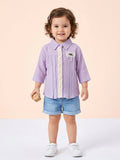 A toddler girl with curly hair wearing a lilac purple muslin long-sleeve shirt with vertical pleats, a central lace trim, and a decorative pearl brooch accent.