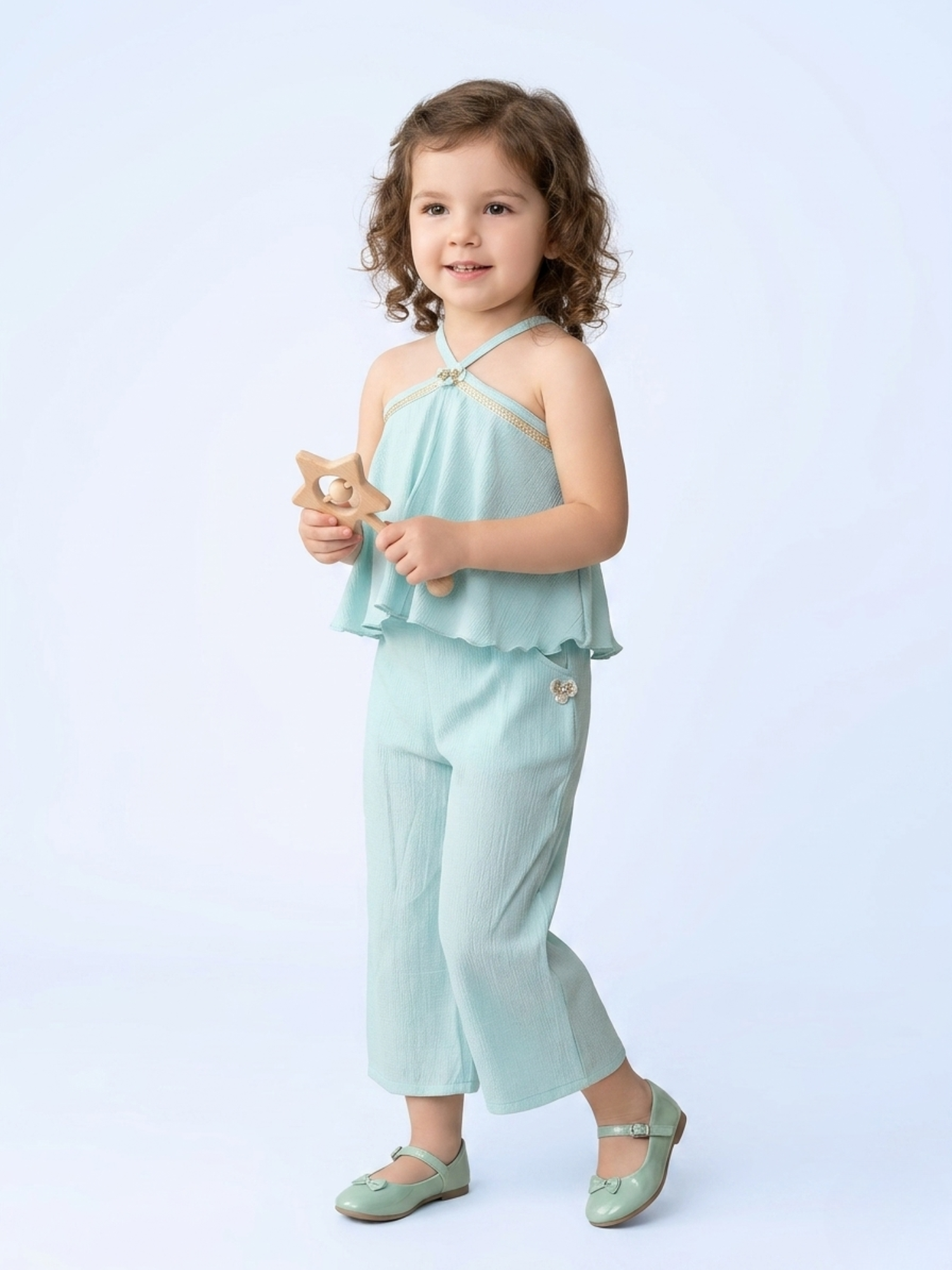 In the image titled "Untitled design(19).png", a toddler girl with curly hair stands smiling while wearing a refreshing mint green co-ord set. The sleeveless halter-neck top features a double-layered, flared overlay bodice accented with a tiny central motif. It is paired with coordinating wide-leg trousers featuring a subtle, matching floral embellishment on the hip pocket. She is holding a wooden star rattle against a soft, plain light blue background.