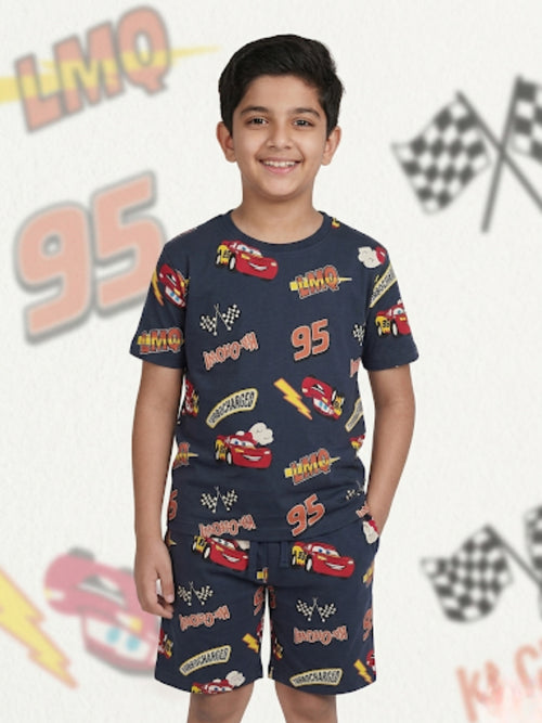 Young boy smiling with hands in pockets wearing a dark grey two-piece cotton set with an all-over Lightning McQueen racing car and checkered flag print.