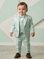 Toddler boy wearing a premium mint green 5-piece tuxedo suit with a patterned jacquard blazer, matching vest, bow tie, and brown formal shoes.
