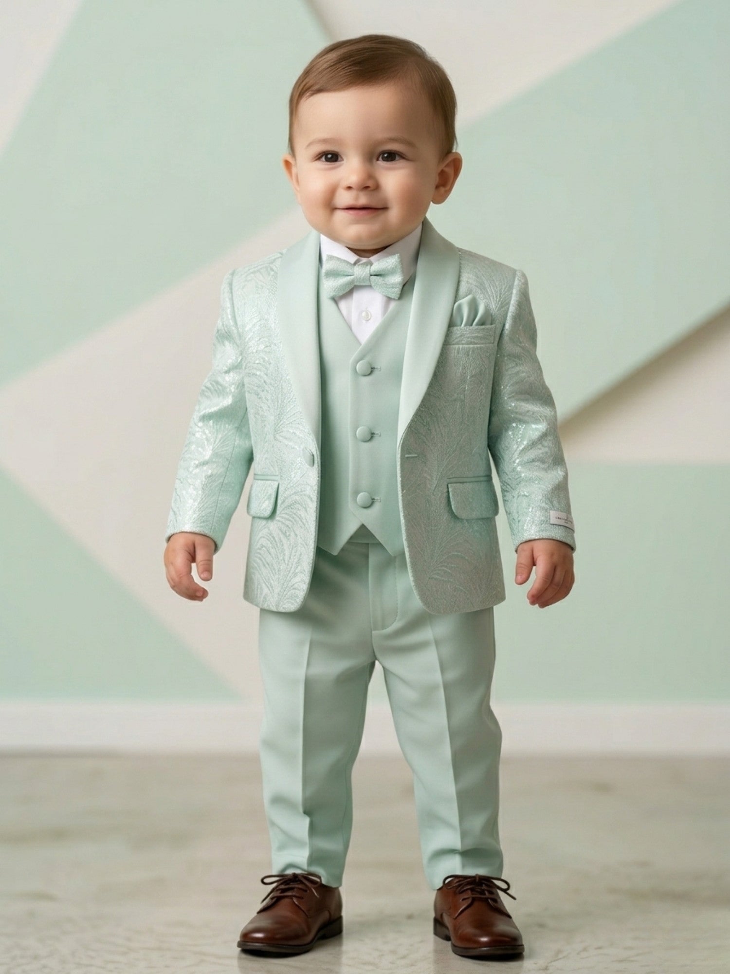 Toddler boy wearing a premium mint green 5-piece tuxedo suit with a patterned jacquard blazer, matching vest, bow tie, and brown formal shoes.