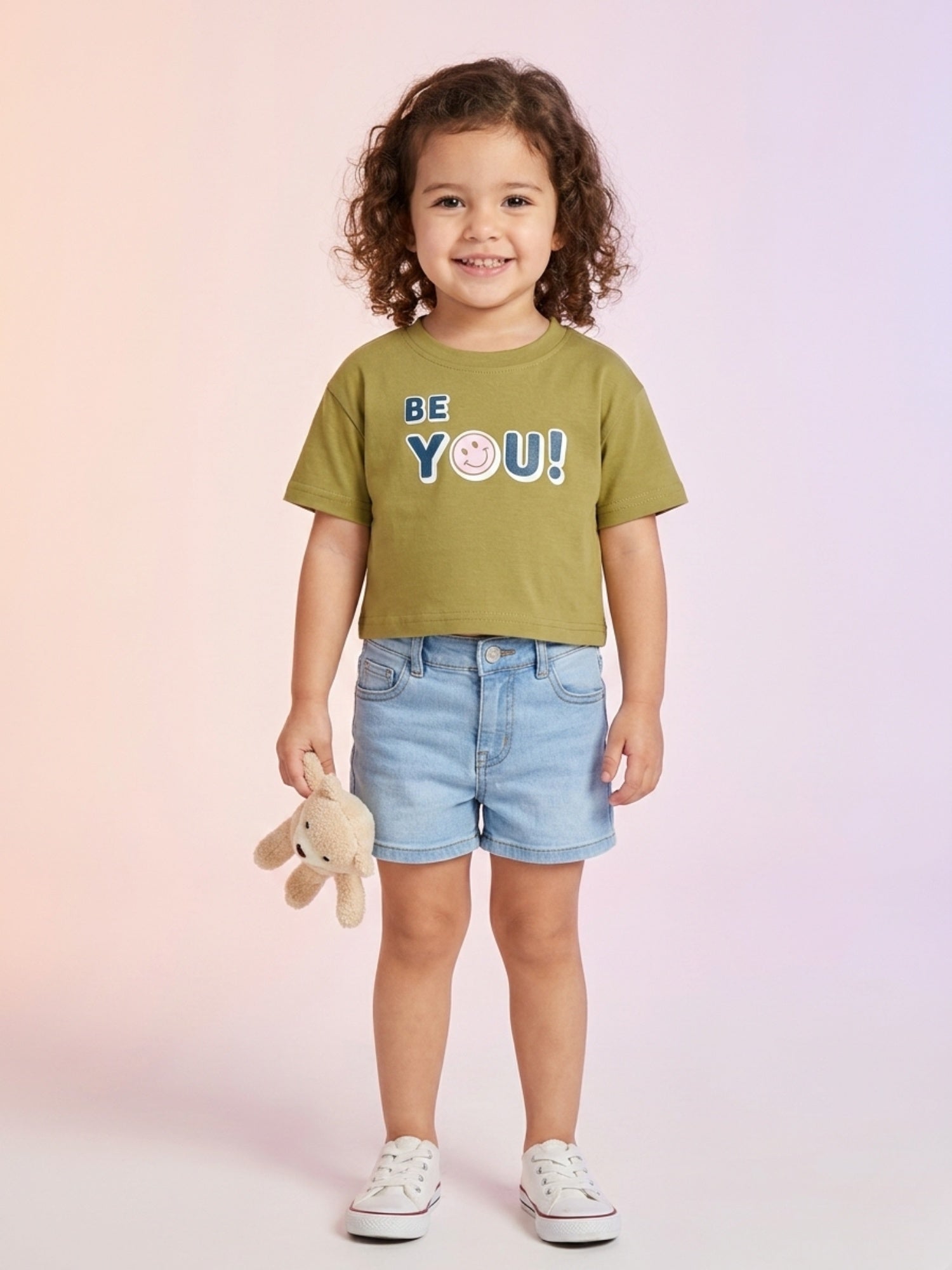 A toddler girl with curly hair wearing an olive green short-sleeve boxy tee with "BE YOU!" and a smiley face graphic, paired with light blue denim shorts.