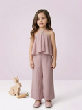 In the image titled "Untitled design(21).png", a young girl stands looking forward while wearing an elegant dusty mauve co-ord set. The sleeveless halter-neck top features a layered, flared overlay bodice accented with a circular pearl and crystal embellishment at the neckline. It is paired with coordinating wide-leg trousers that feature side pockets and a small matching floral applique on the hip. A wooden toy rabbit and carrot rest on the floor next to her against a soft lavender-pink background.