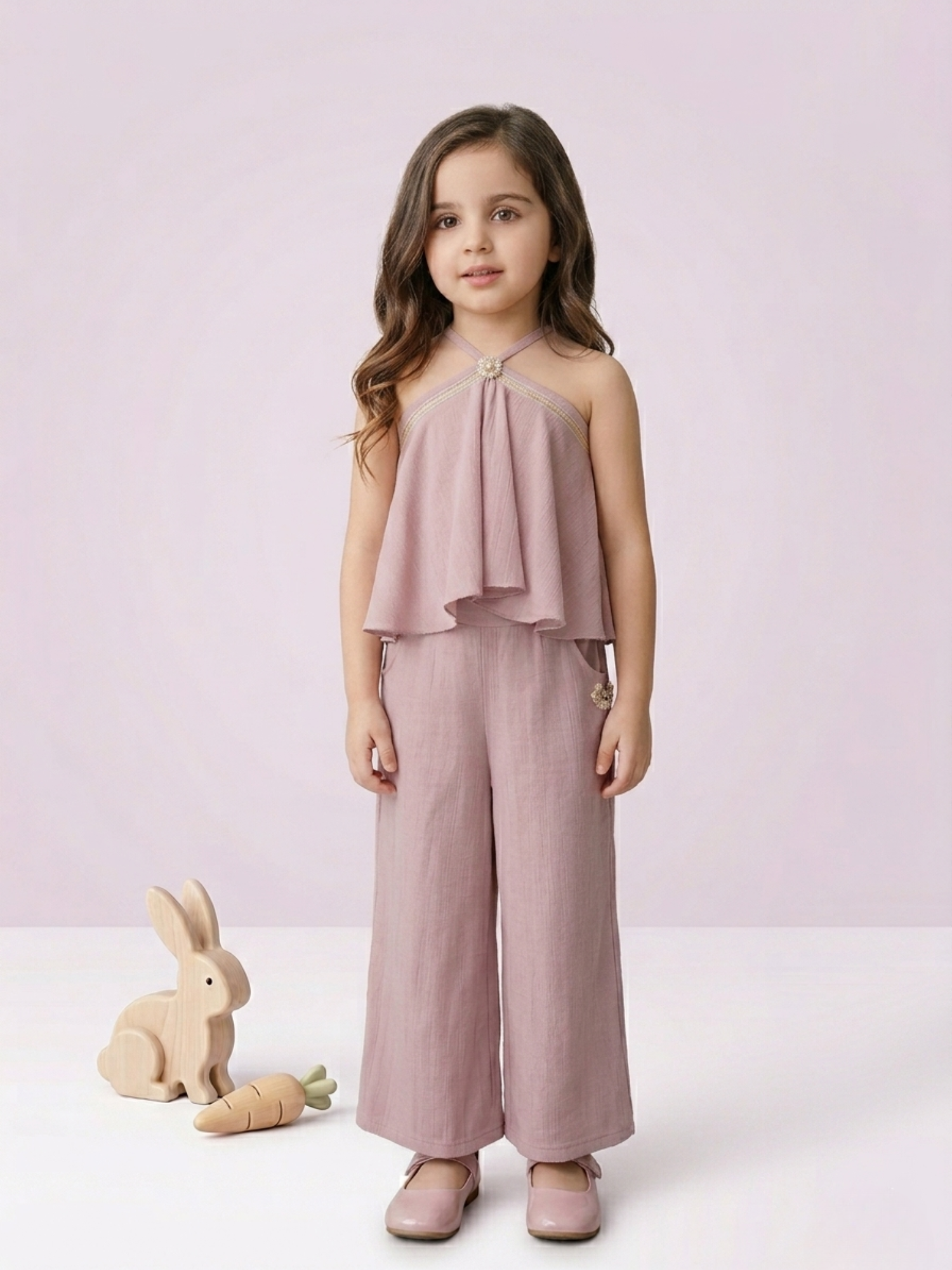 In the image titled "Untitled design(21).png", a young girl stands looking forward while wearing an elegant dusty mauve co-ord set. The sleeveless halter-neck top features a layered, flared overlay bodice accented with a circular pearl and crystal embellishment at the neckline. It is paired with coordinating wide-leg trousers that feature side pockets and a small matching floral applique on the hip. A wooden toy rabbit and carrot rest on the floor next to her against a soft lavender-pink background.