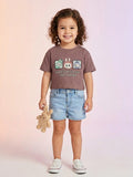A toddler girl with curly hair wearing a cocoa brown short-sleeve boxy tee featuring Labubu characters and the text "Cuteness with a bite. That's Labubu," paired with light-wash denim shorts.