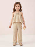In the image titled "Untitled design(22).png", a young girl stands smiling while wearing a chic beige linen halter-neck co-ord set. The sleeveless top features a tiered flare design, delicate cream lace trim, and a small textured bow applique at the neckline. The matching straight-leg trousers include side pockets and a coordinating bow detail on the left hip. She holds a small wooden star toy against a soft, neutral beige background.