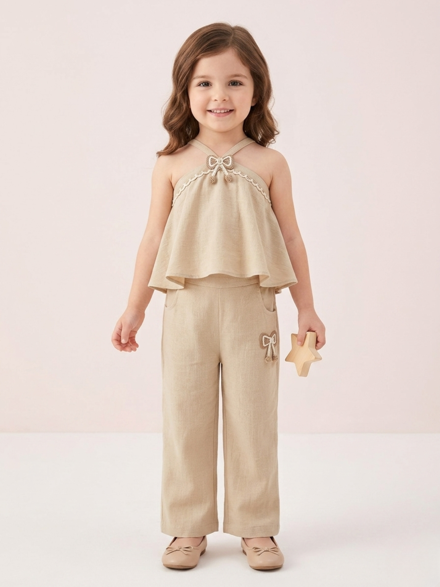 In the image titled "Untitled design(22).png", a young girl stands smiling while wearing a chic beige linen halter-neck co-ord set. The sleeveless top features a tiered flare design, delicate cream lace trim, and a small textured bow applique at the neckline. The matching straight-leg trousers include side pockets and a coordinating bow detail on the left hip. She holds a small wooden star toy against a soft, neutral beige background.