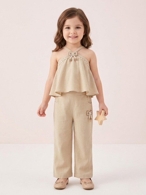 In the image titled "Untitled design(22).png", a young girl stands smiling while wearing a chic beige linen halter-neck co-ord set. The sleeveless top features a tiered flare design, delicate cream lace trim, and a small textured bow applique at the neckline. The matching straight-leg trousers include side pockets and a coordinating bow detail on the left hip. She holds a small wooden star toy against a soft, neutral beige background.