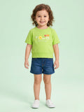 A smiling toddler girl with curly hair wearing a vibrant lime green short-sleeve tee with "FLOWER" and a small floral graphic on the front, paired with cuffed denim shorts.