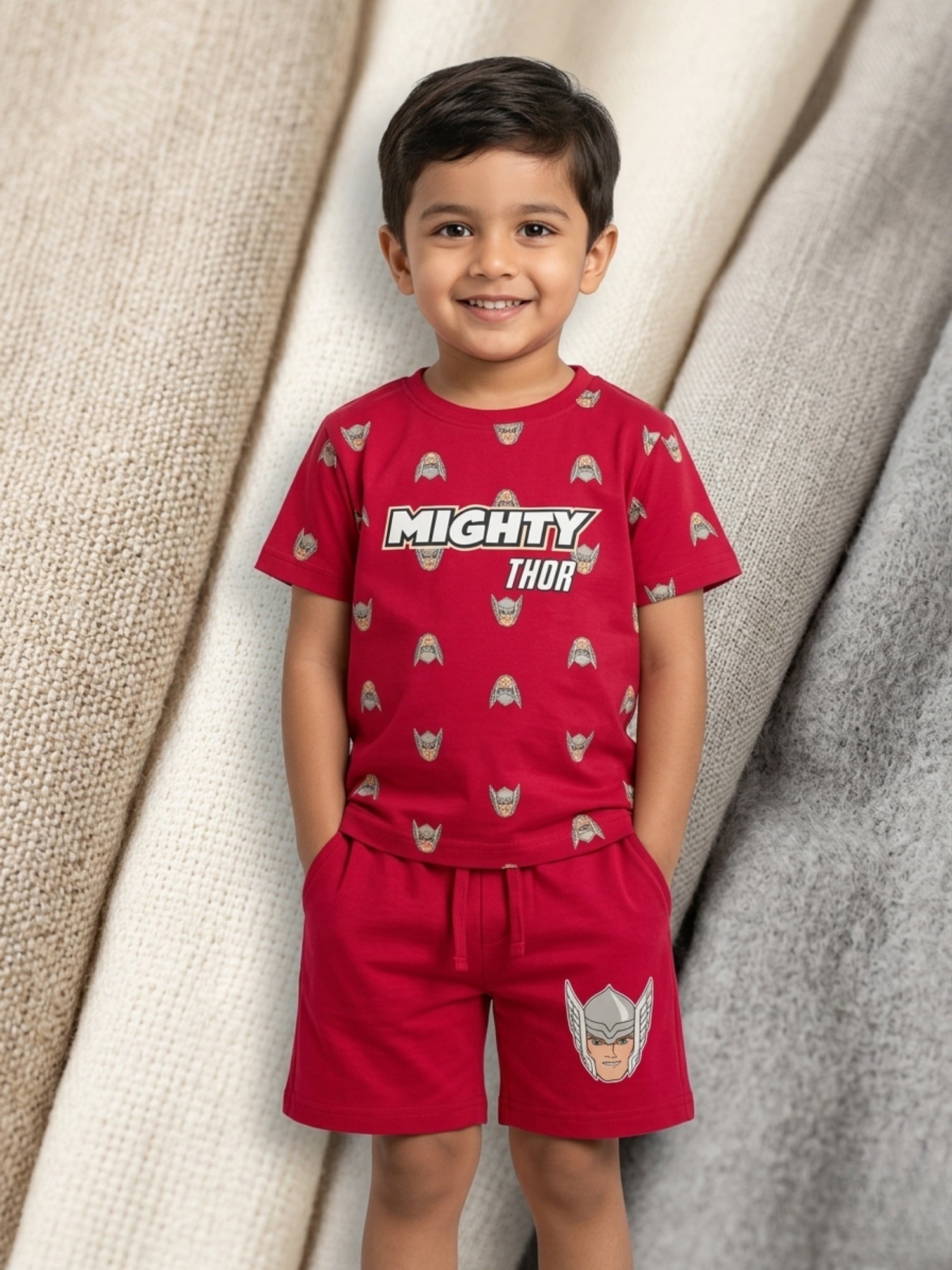 Young boy smiling with hands in pockets wearing a bright red two-piece cotton set with an all-over Mighty Thor helmet and text print.