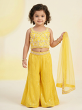 A young girl smiling in a bright yellow sleeveless choli with floral embroidery and matching sequined sharara pants, holding a sheer yellow dupatta.