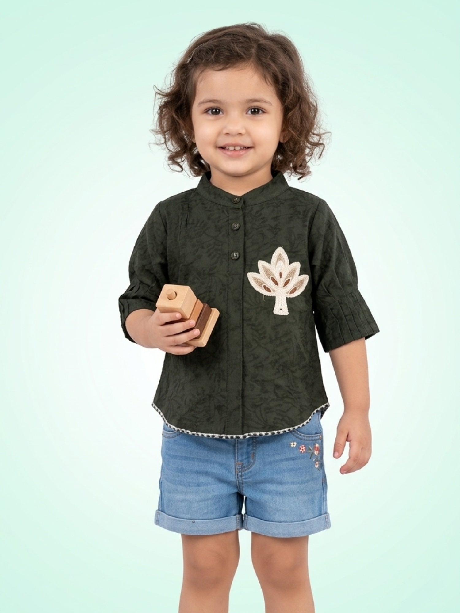 Young girl wearing an olive green mandarin collar shirt with a large leaf applique and a delicate scalloped bottom edge.
