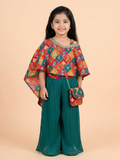 A young girl modeling a vibrant multicolor asymmetrical patchwork poncho top with sequins and mirror work, paired with teal green pleated wide-leg palazzo pants and a matching mini purse.