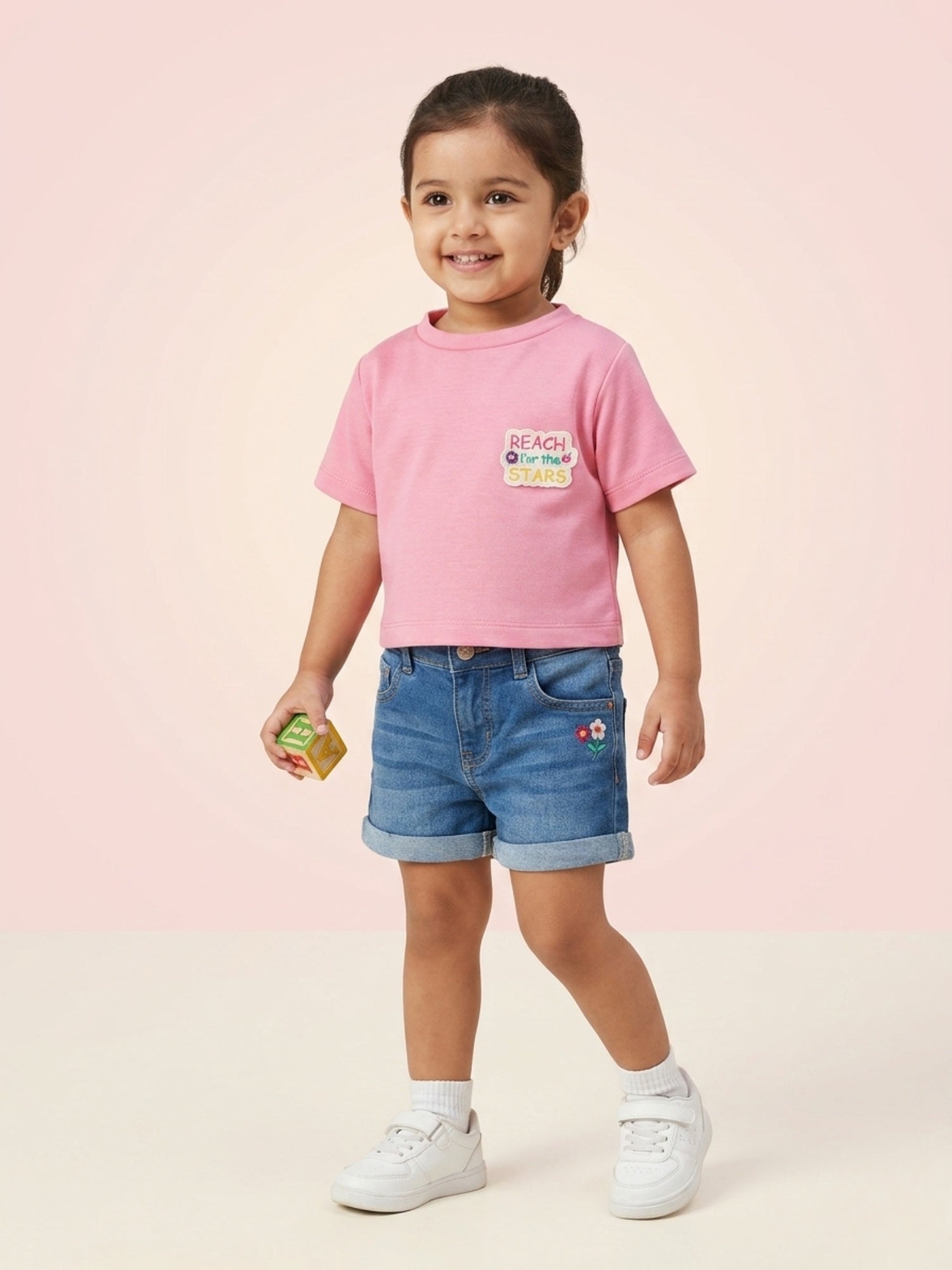 A young girl wearing a bright bubblegum pink short-sleeve tee with a "Reach for the Stars" patch on the chest, paired with embroidered denim shorts.