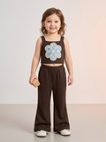 A young girl standing in a studio setting wearing a two-piece casual set from Kiya Store, featuring a deep brown sleeveless crop top with a large blue and white embroidered flower on the front, paired with matching brown wide-leg flared pants.