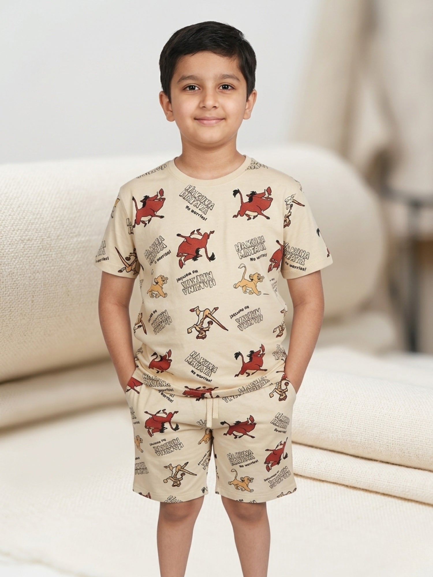Young boy with hands in pockets wearing a beige two-piece cotton set with an all-over Lion King character print featuring Simba and Pumbaa.
