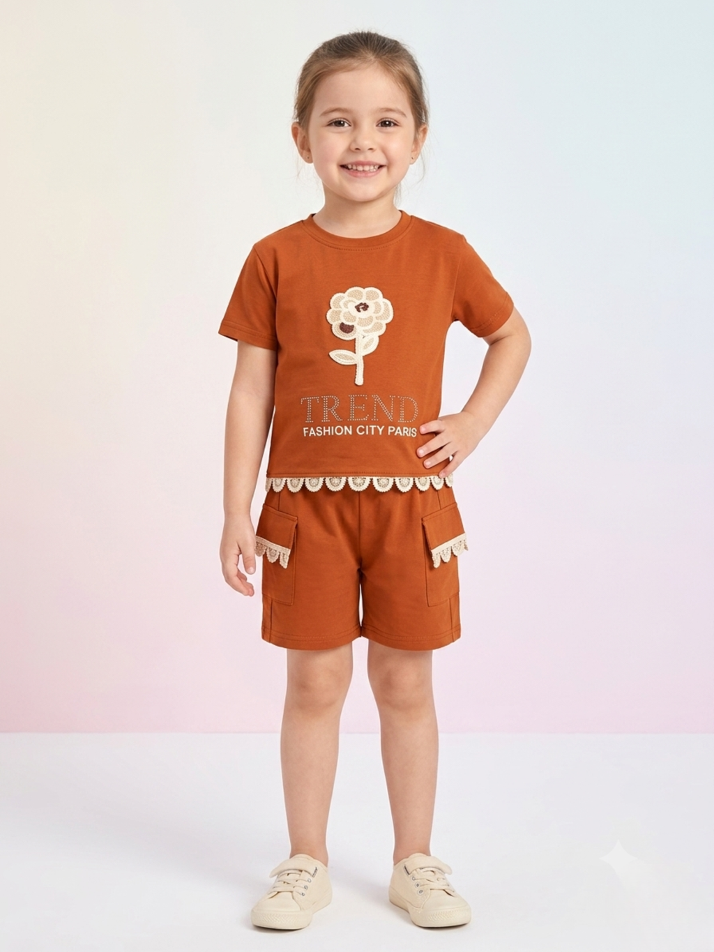 a smiling young girl wears a rust-brown short-sleeve t-shirt with a crochet flower appliqué and "TREND" lettering, paired with matching rust cargo shorts trimmed in scalloped lace.