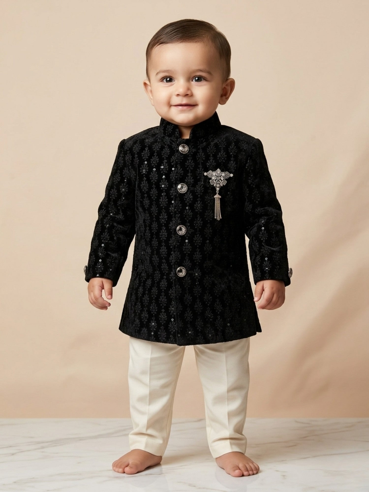 Baby boy wearing black embroidered ethnic kurta with cream pants and decorative brooch for festive and wedding occasions.
