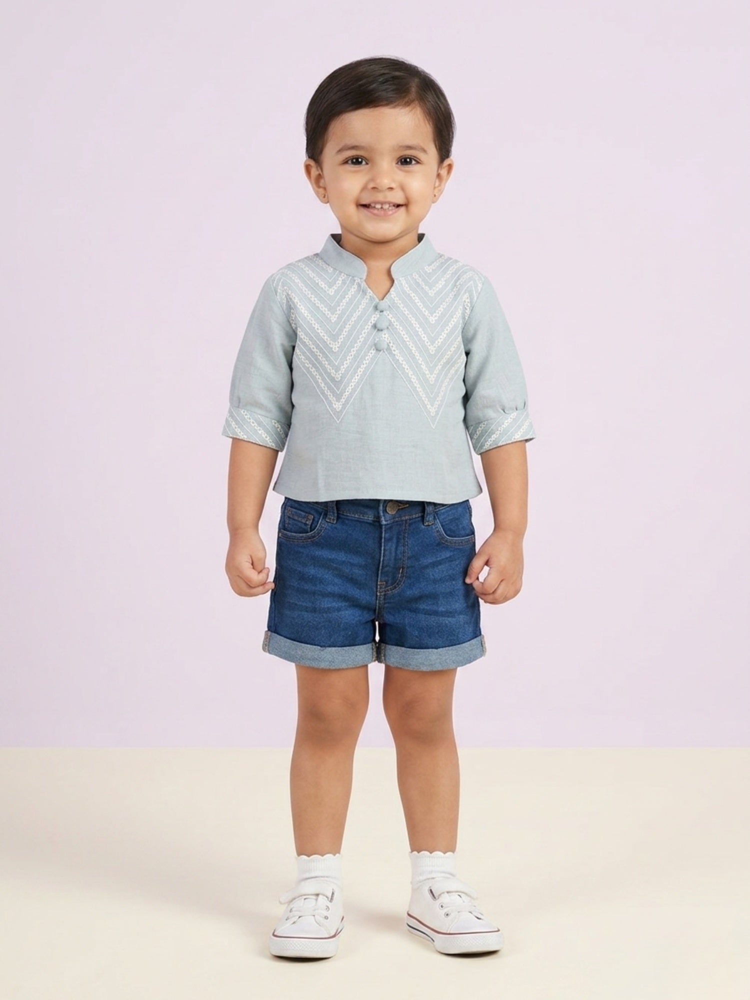 A toddler girl with curly hair wearing a deep teal short-sleeve button-up top with white vertical lace trim and a matching white lace collar.