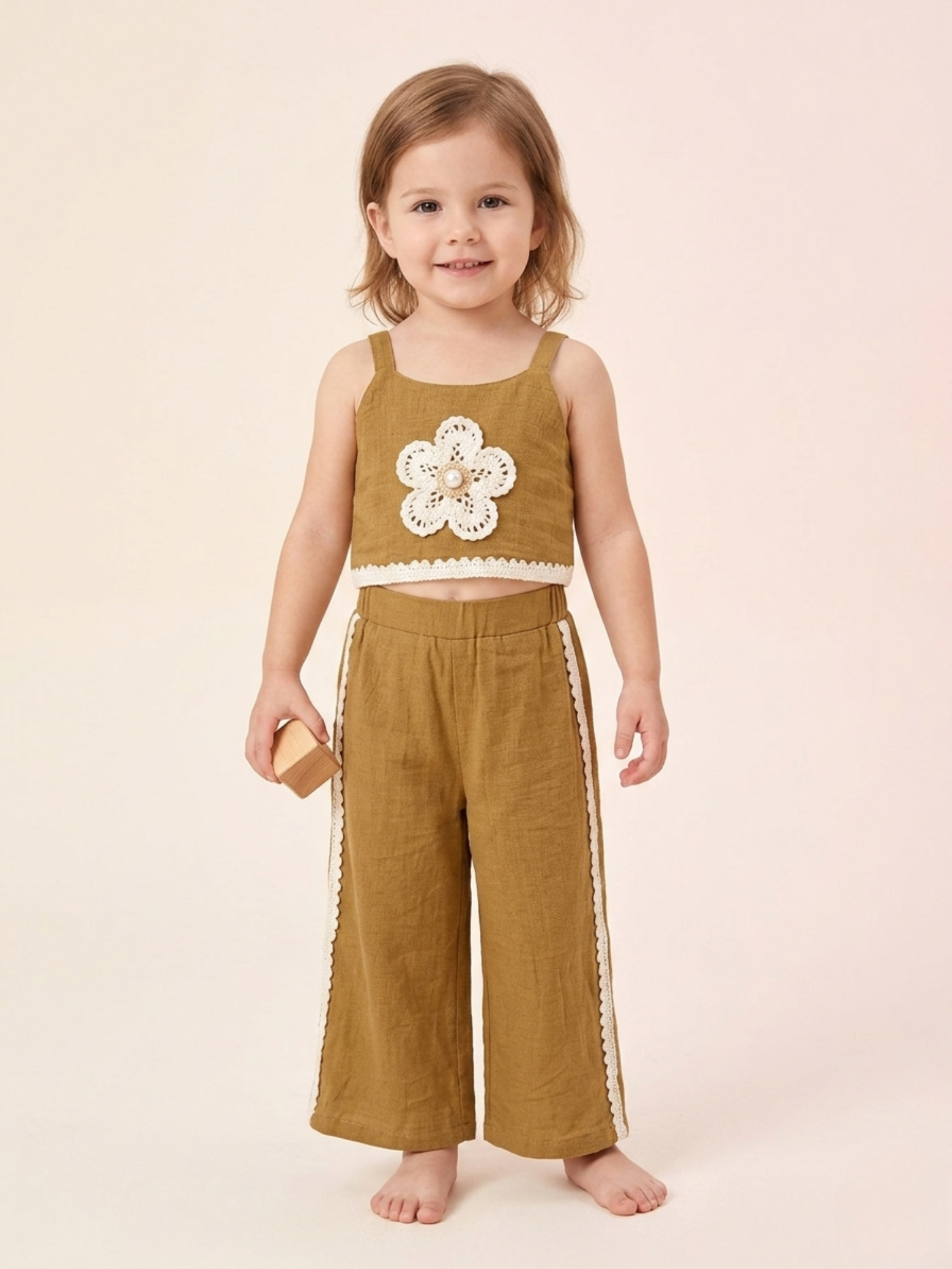 A young girl standing barefoot in a studio setting wearing a two-piece linen set from Kiya Store, featuring a mustard yellow sleeveless crop top with a large crochet flower and white lace hem, paired with matching wide-leg pants with white lace side panels.
