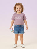 A toddler girl with curly hair smiling and wearing a mauve-purple short-sleeve boxy tee with "SUNDAY" printed in a white frame, paired with denim shorts.