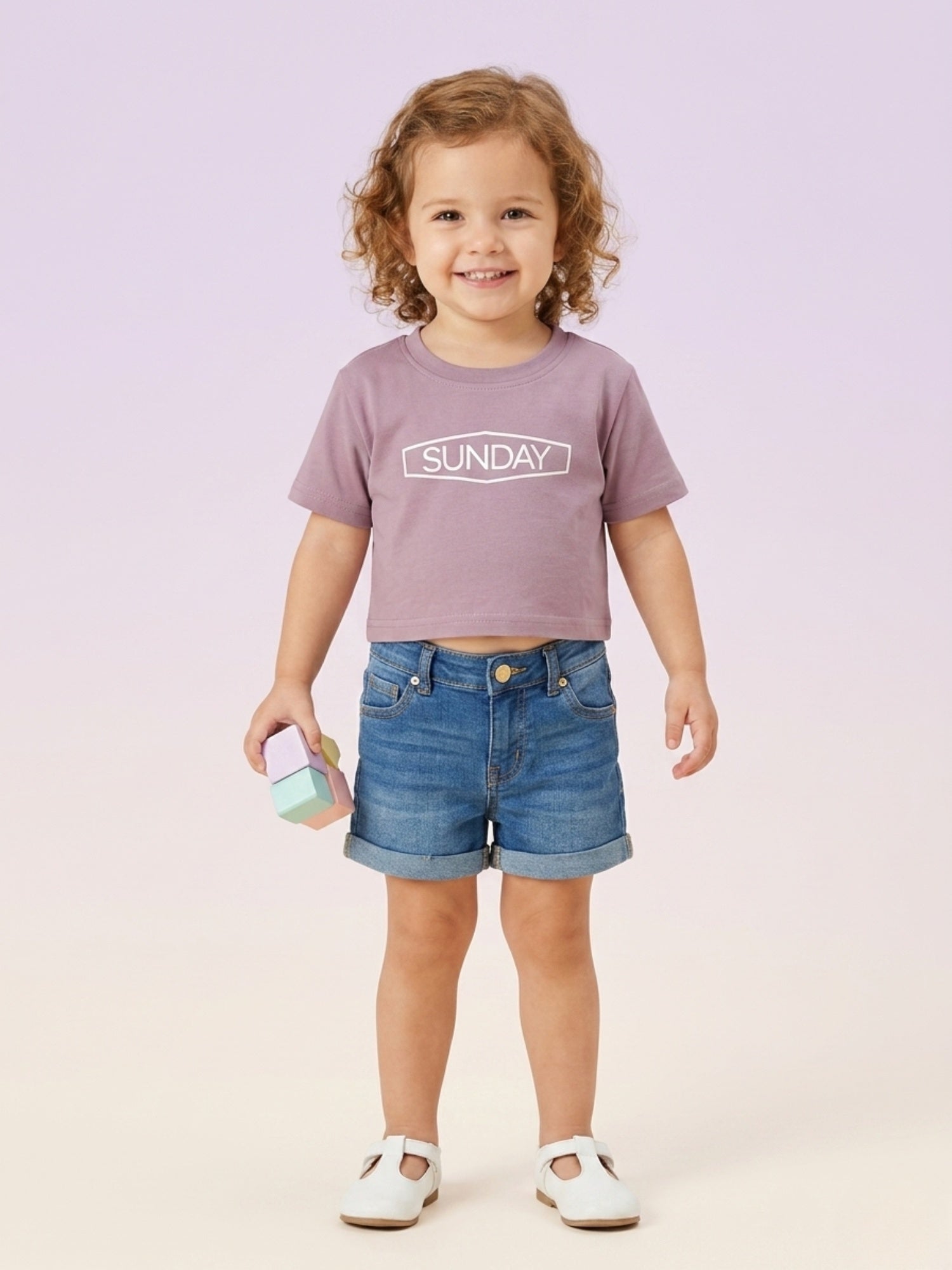 A toddler girl with curly hair smiling and wearing a mauve-purple short-sleeve boxy tee with "SUNDAY" printed in a white frame, paired with denim shorts.