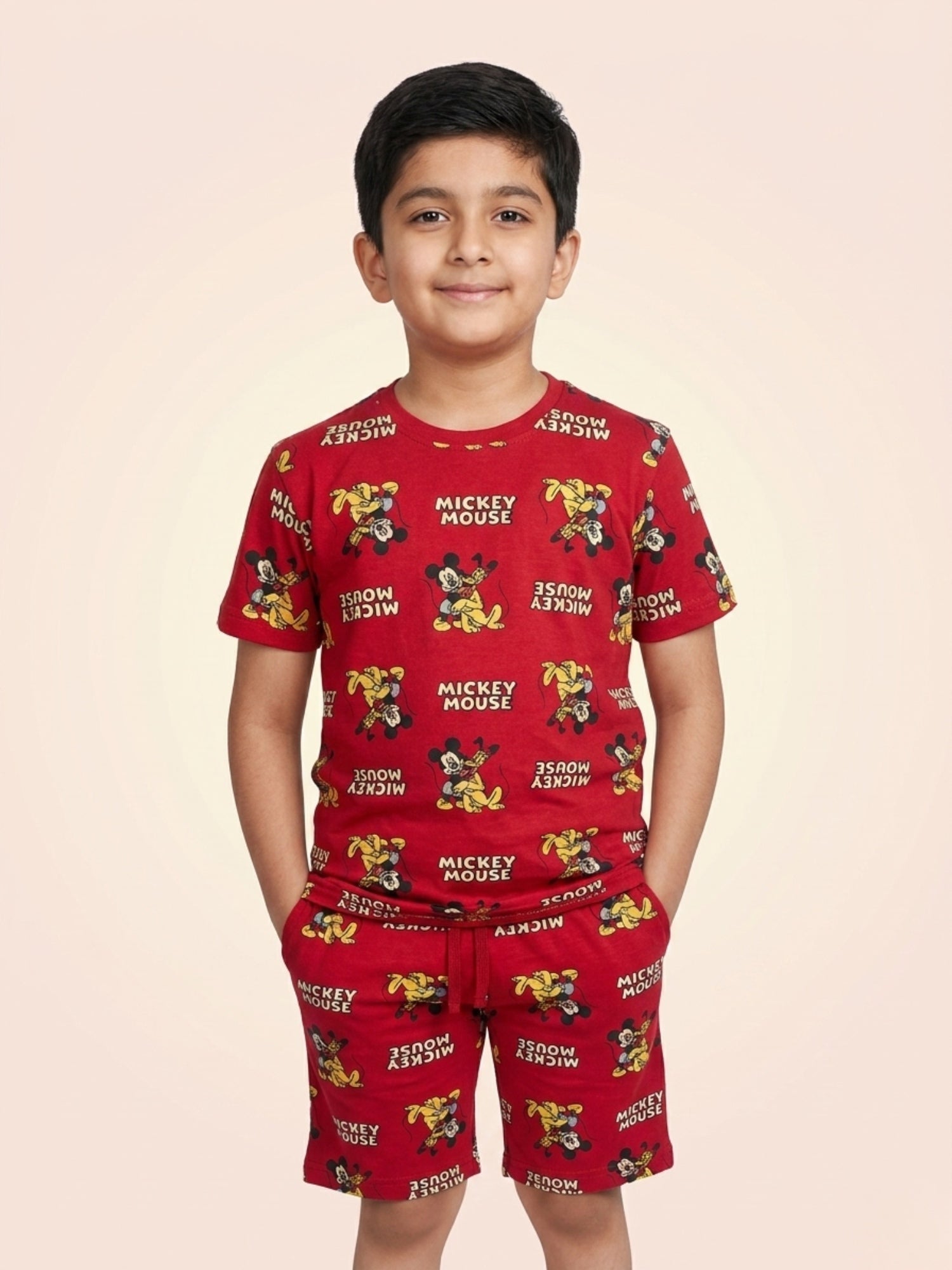 Young boy smiling with hands in pockets wearing a bright red two-piece cotton set with an all-over Mickey Mouse character and text print.
