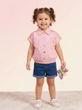 A smiling toddler girl with pigtails wearing a short-sleeve pink button-down shirt featuring colorful rainbow stripes on the shoulders, multi-colored buttons, and pink floral appliqué patches.