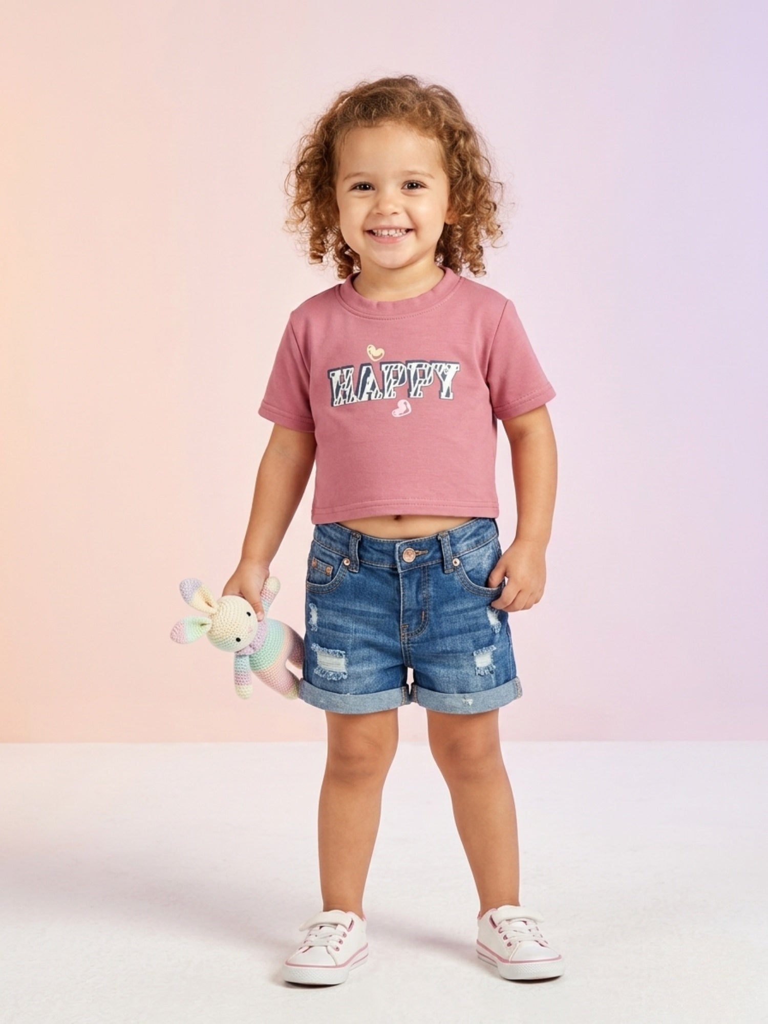 A smiling toddler girl with curly hair wearing a dusty rose short-sleeve crop-style tee with "HAPPY" printed on the front, paired with distressed denim shorts.