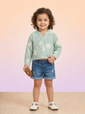 A toddler girl with curly hair wearing a mint green muslin long-sleeve top with white floral eyelet embroidery, paired with denim shorts and white t-strap shoes.
