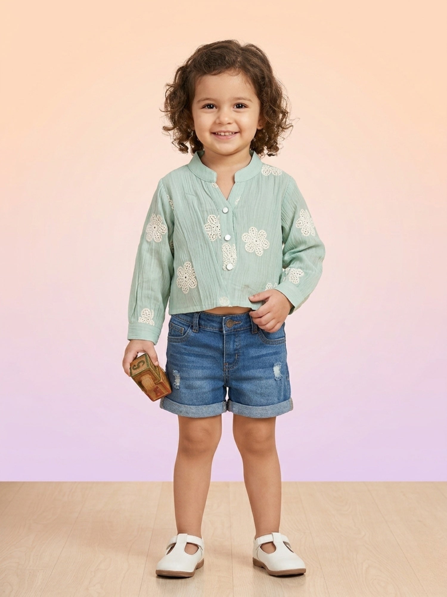 A toddler girl with curly hair wearing a mint green muslin long-sleeve top with white floral eyelet embroidery, paired with denim shorts and white t-strap shoes.