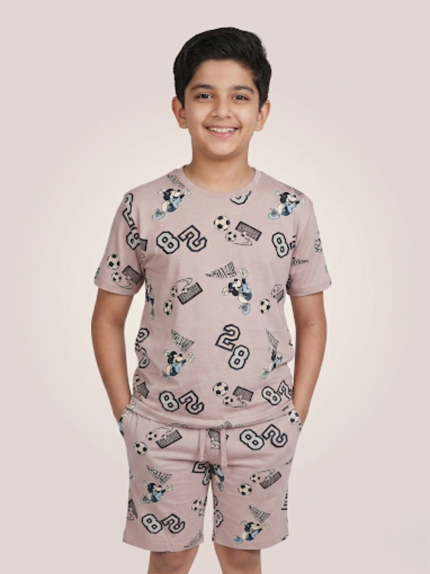 Young boy smiling with hands in pockets wearing a mauve two-piece cotton set with an all-over football-themed Mickey Mouse and soccer ball print.